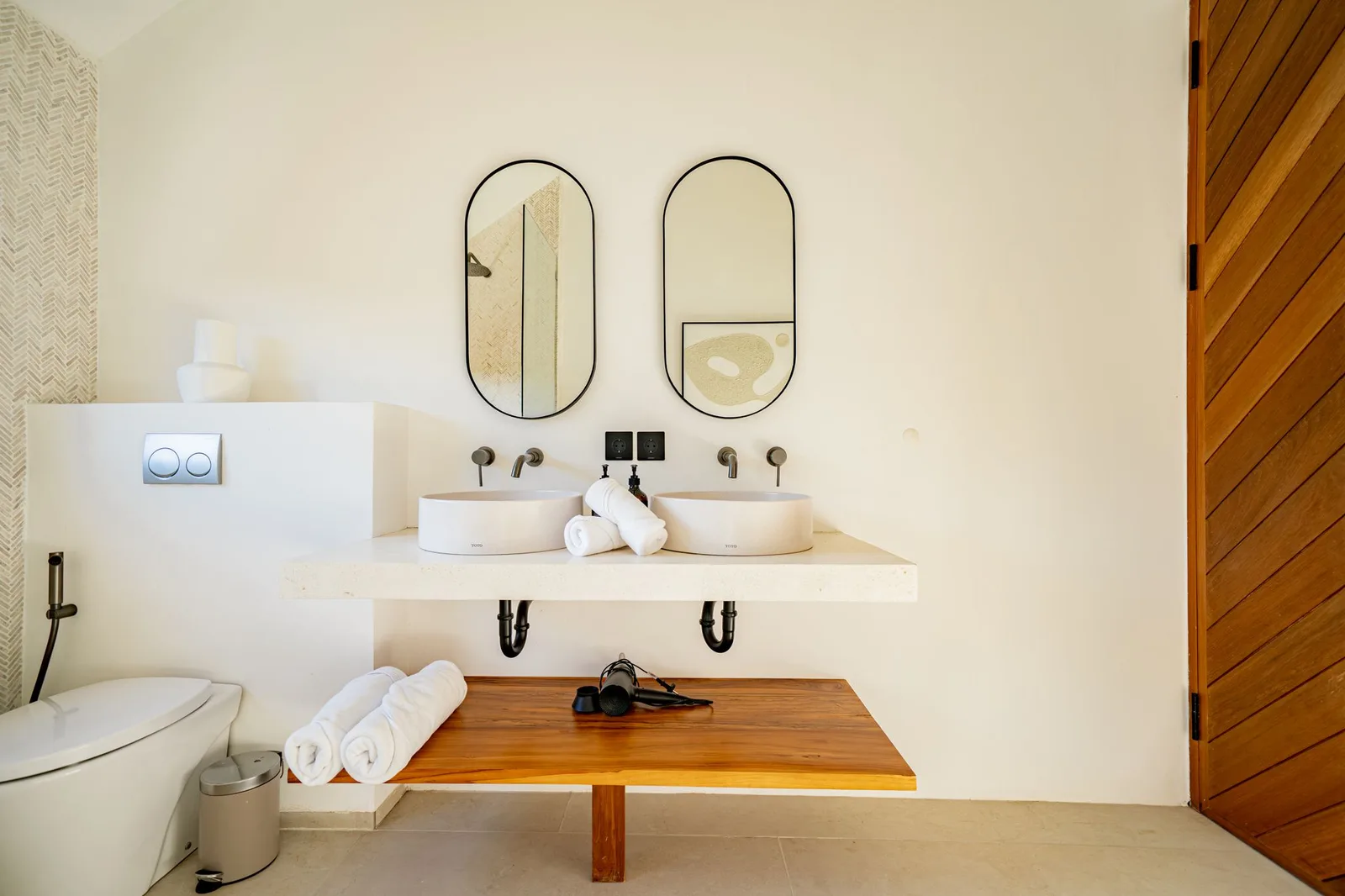 A bright bathroom has a double sink vanity, two arched mirrors, a toilet, and a wooden countertop.