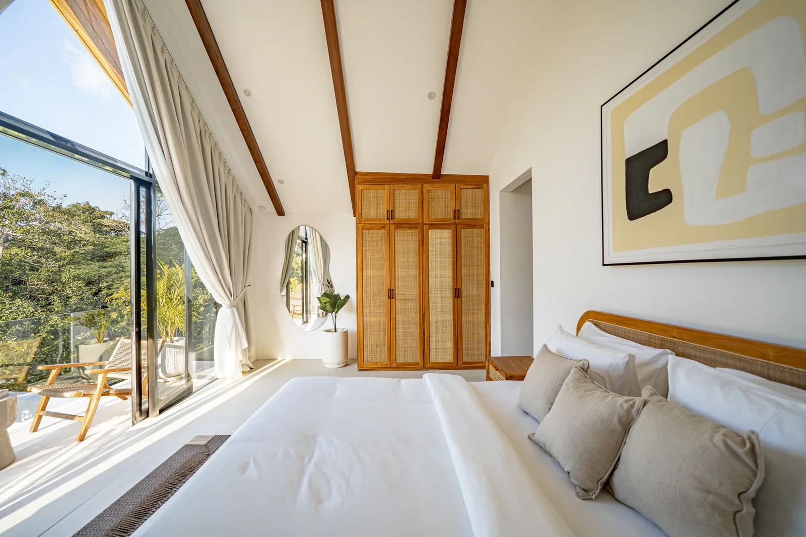 A bright bedroom has a white bed, a wooden louvered wardrobe, and glass doors opening to a balcony.