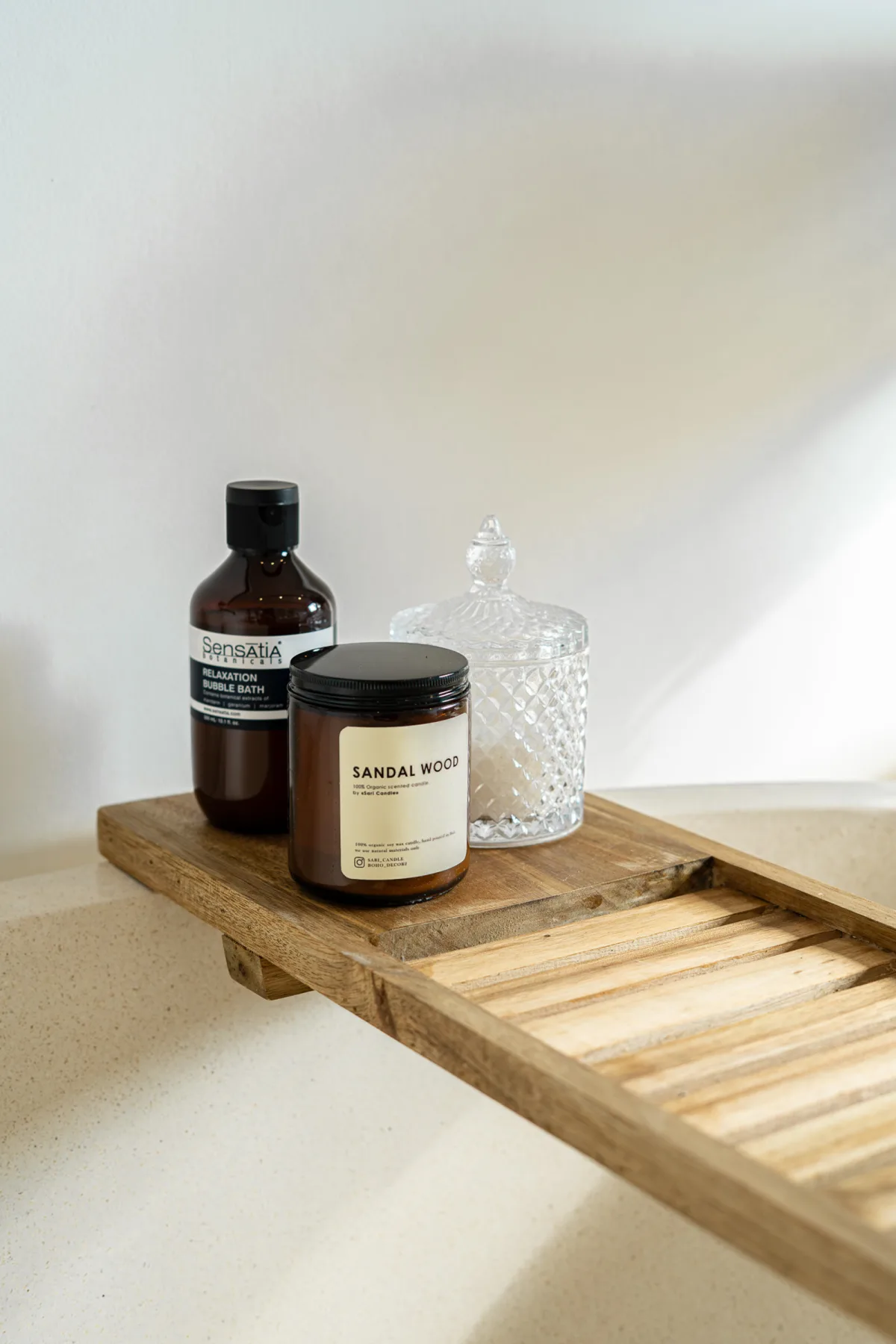 A wooden bath tray holds a dark bottle, a sandalwood candle, and a clear glass jar.