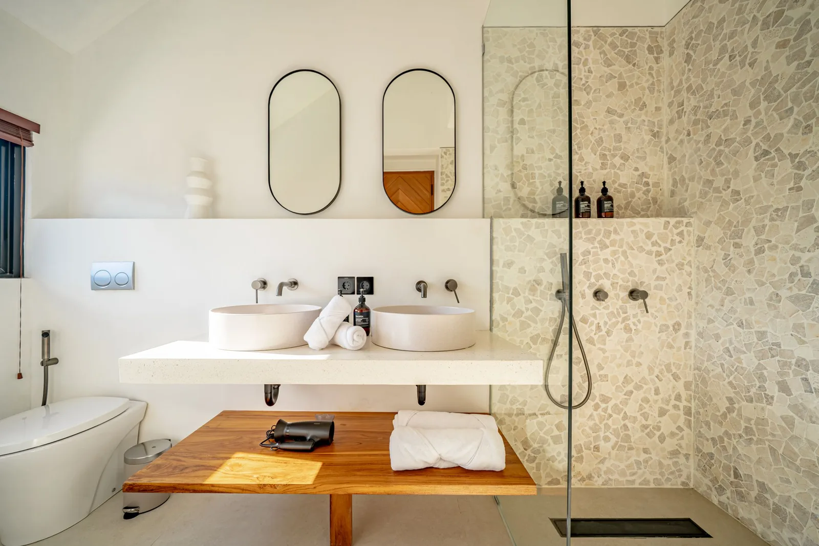 A bright bathroom has a double-sink vanity, a wooden bench, and a glass-enclosed stone shower.