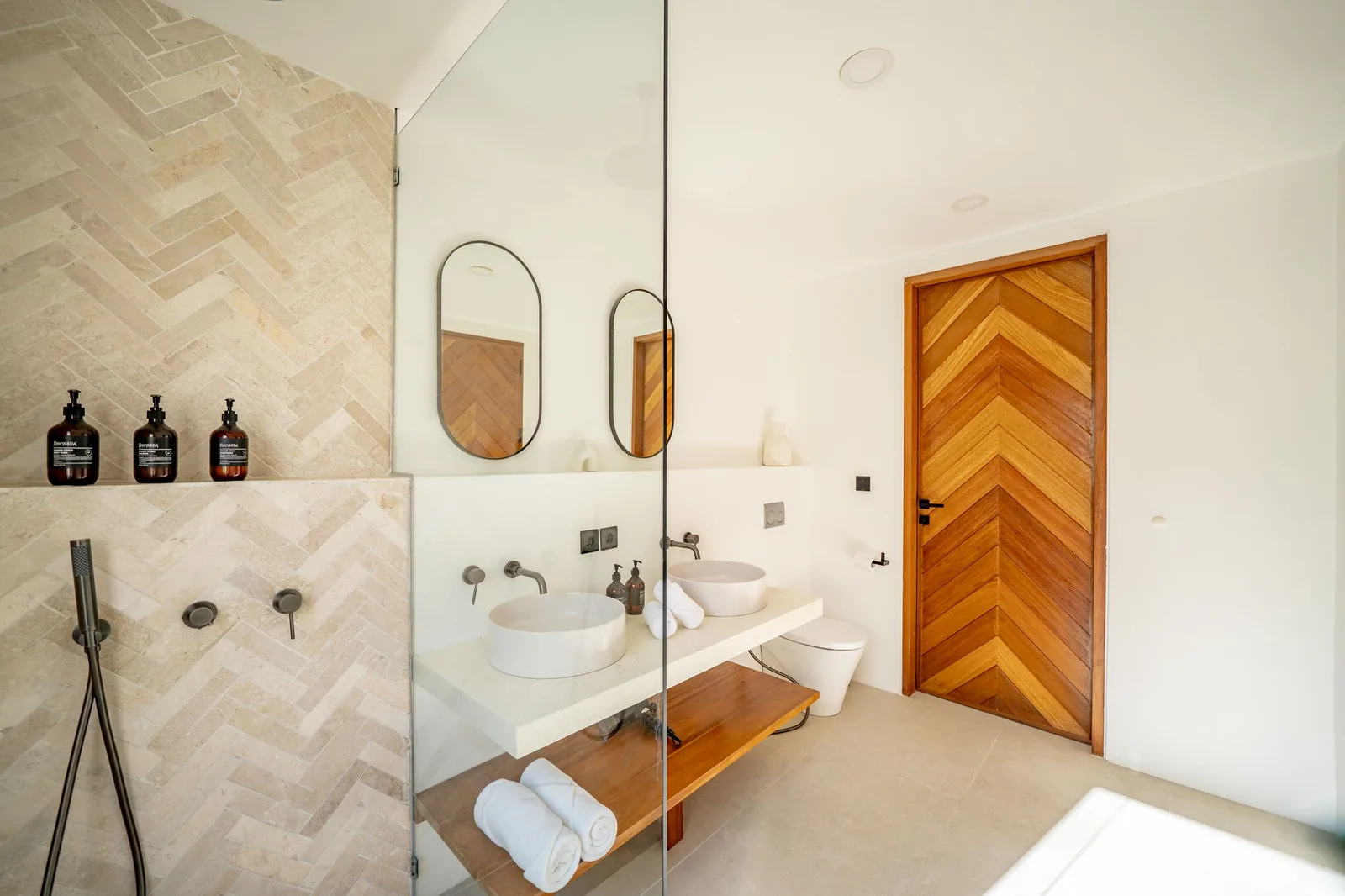 A bright bathroom with a glass shower, twin vessel sinks, wall mirrors, and a wooden chevron-pattern door.