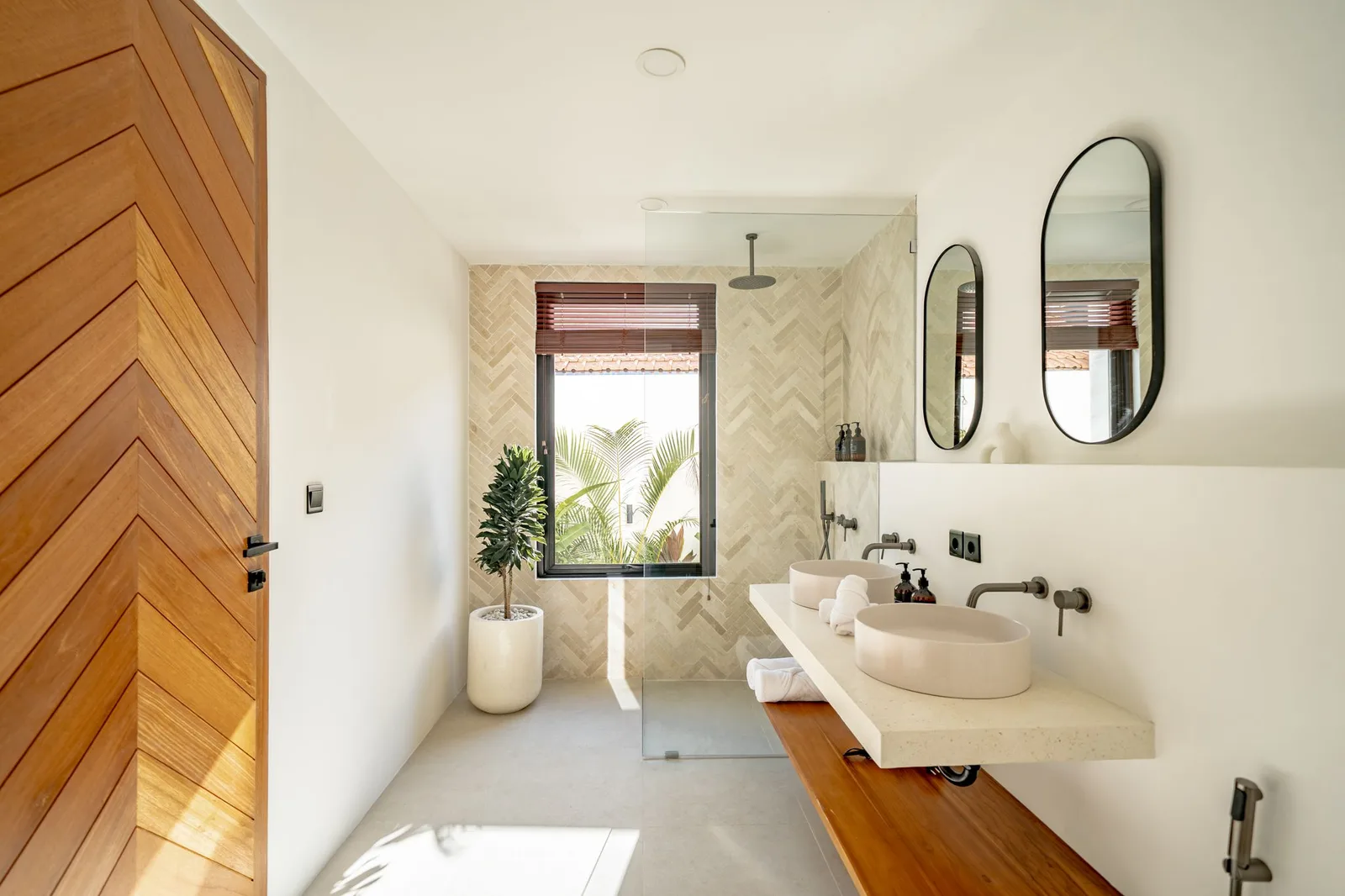 A modern bathroom features a walk-in shower, a vessel sink, oval mirrors, and a window with a woven blind.