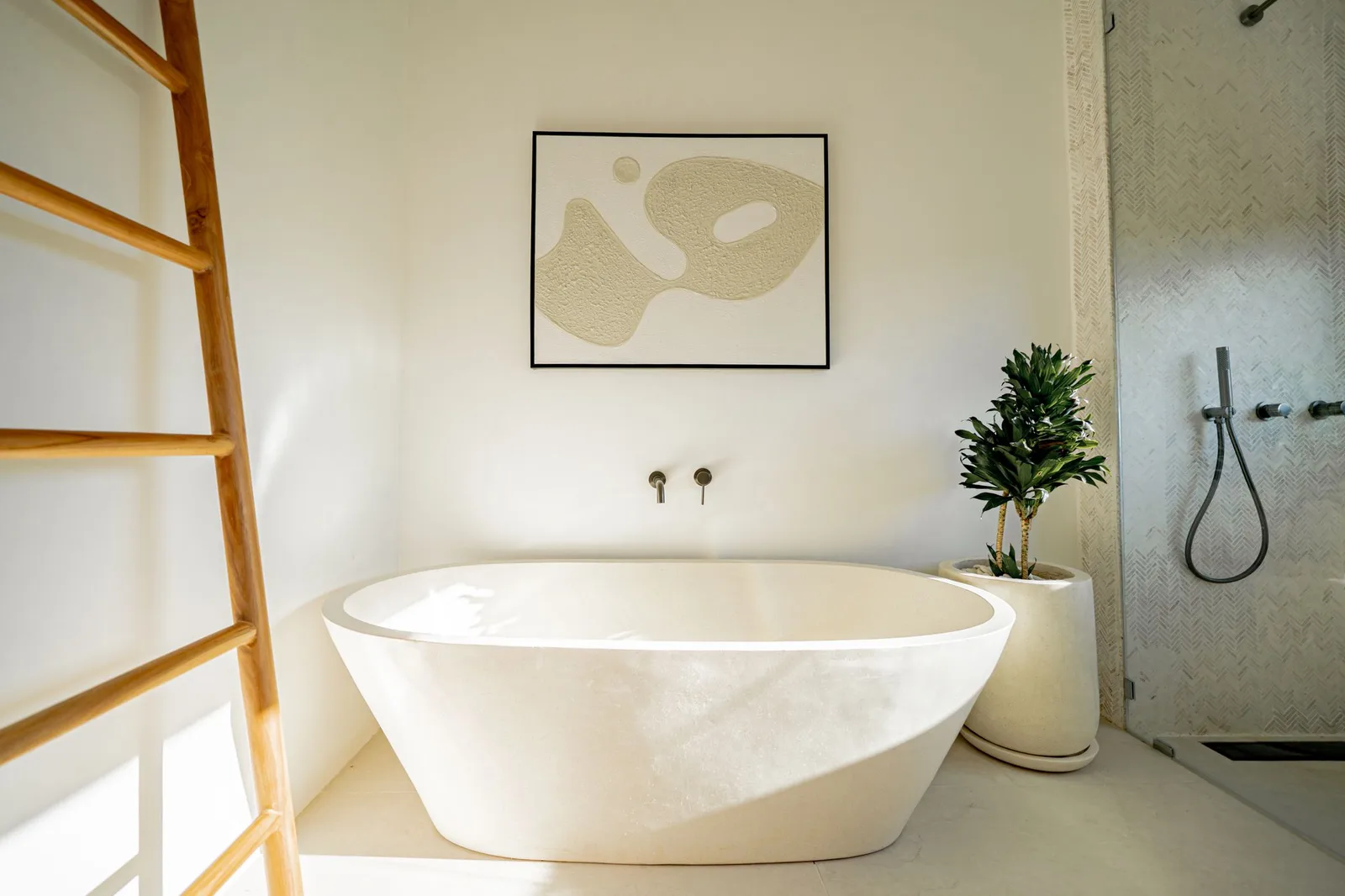A freestanding white bathtub sits beside a glass shower, with a wooden ladder and potted plant.