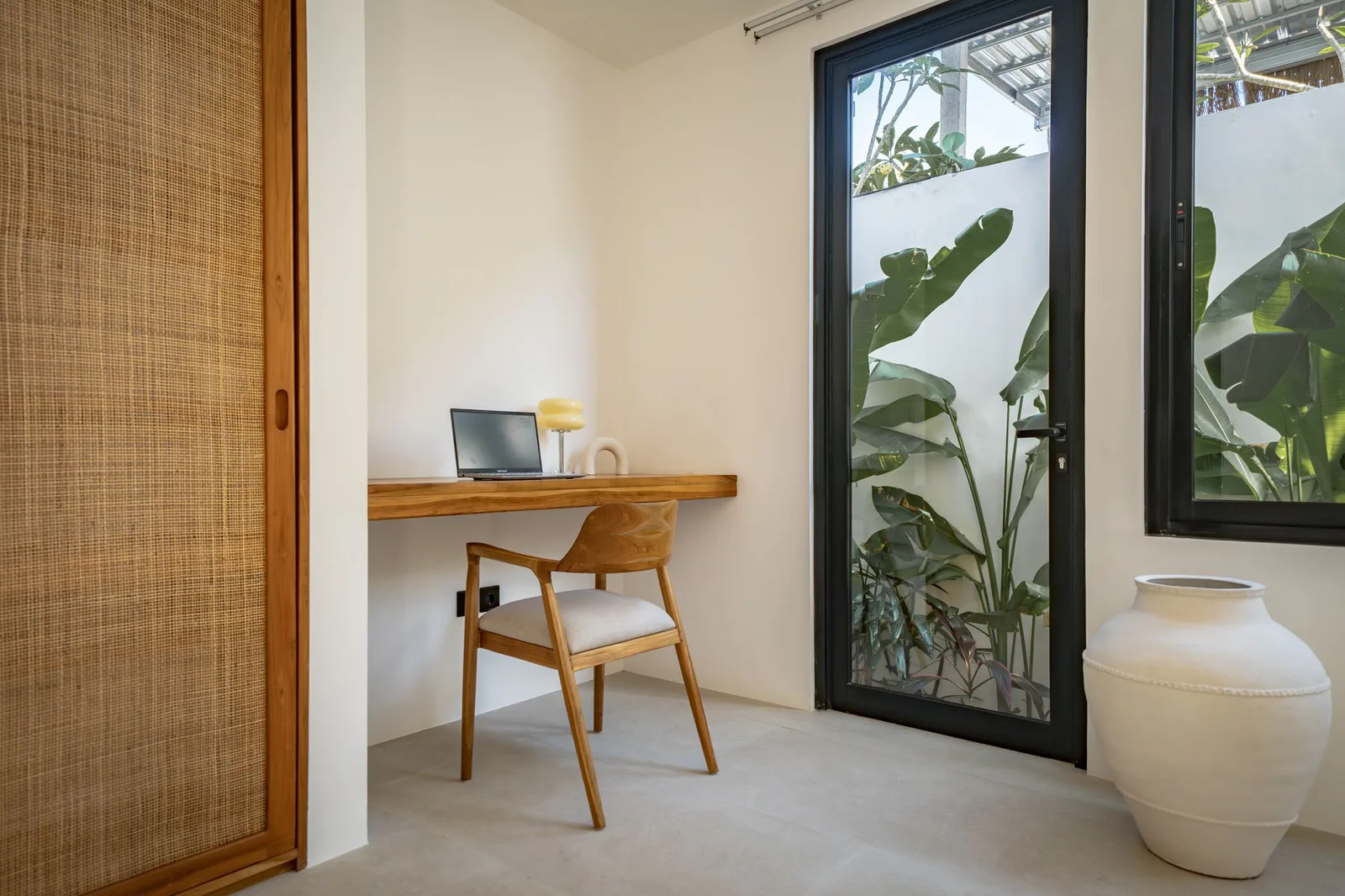 A bright room has a small wooden desk and chair beside a glass door overlooking leafy plants.