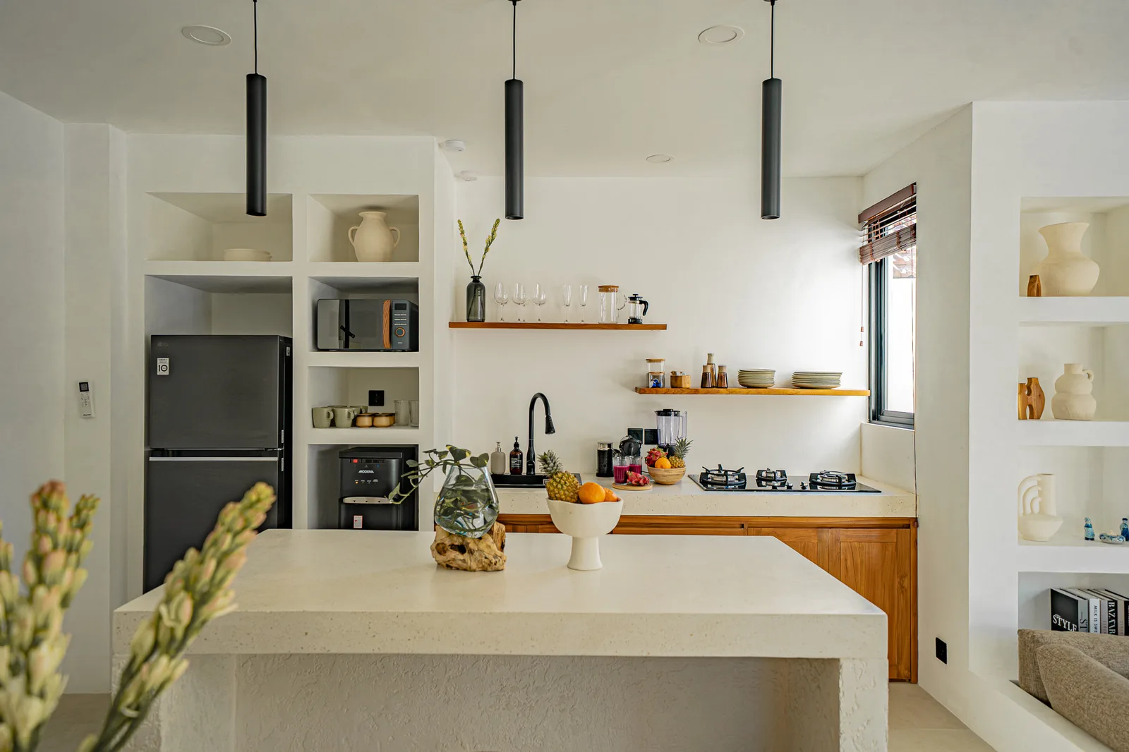 A bright modern kitchen features a large stone island, built-in appliances, open shelves, and pendant lights.