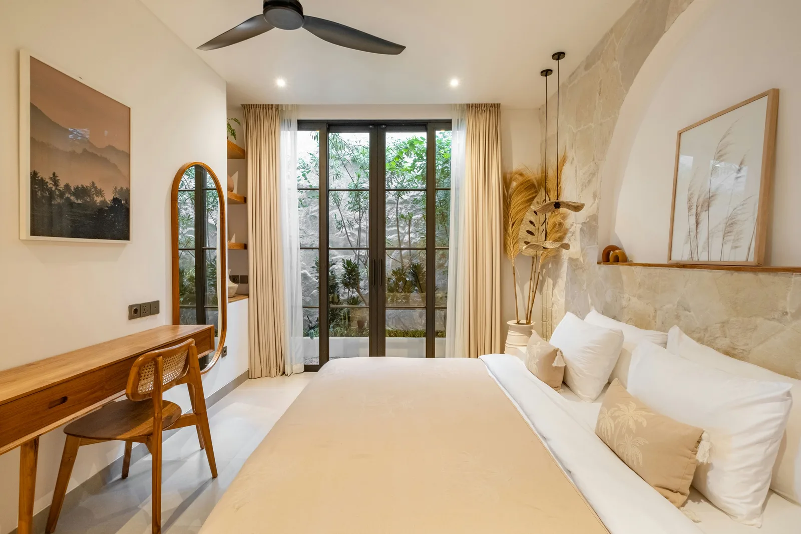Un dormitorio luminoso cuenta con una cama grande, un escritorio de madera y puertas de vidrio hacia una zona exterior frondosa.