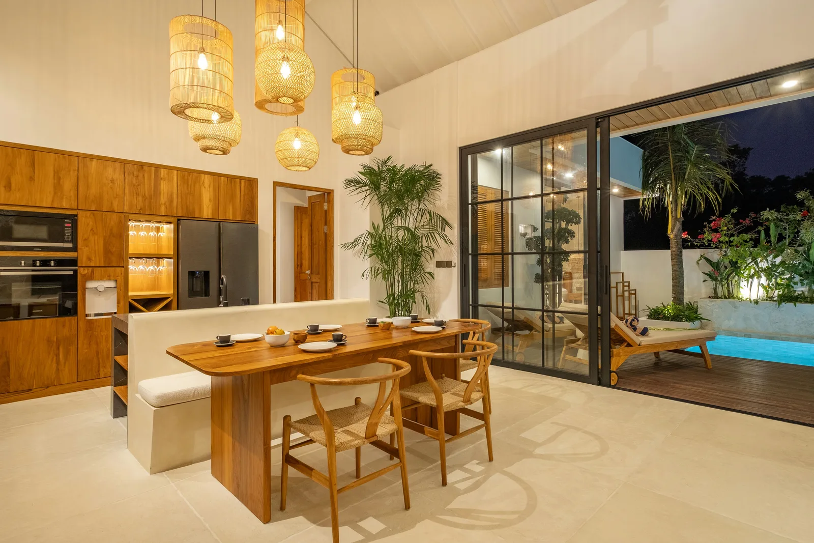 Un luminoso comedor de planta abierta tiene una mesa de madera, lámparas colgantes tejidas y puertas corredizas de vidrio hacia un patio.