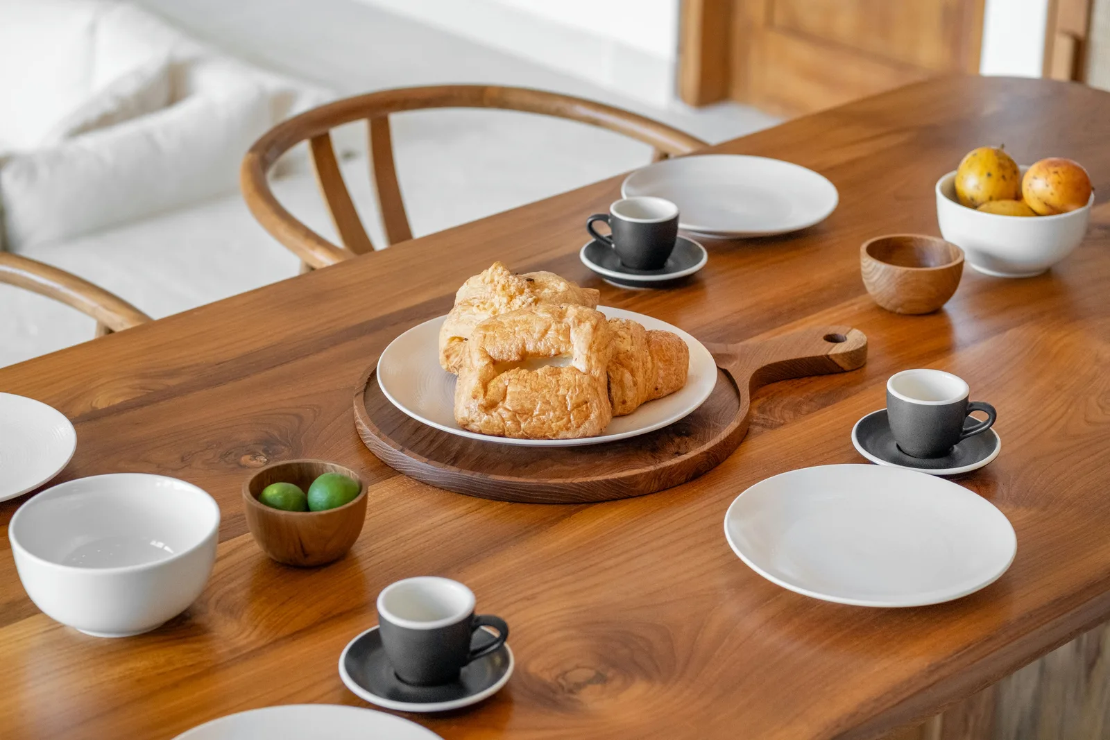 Una mesa de comedor de madera está dispuesta con bollería, platos blancos, tazas de café y cuencos de fruta.