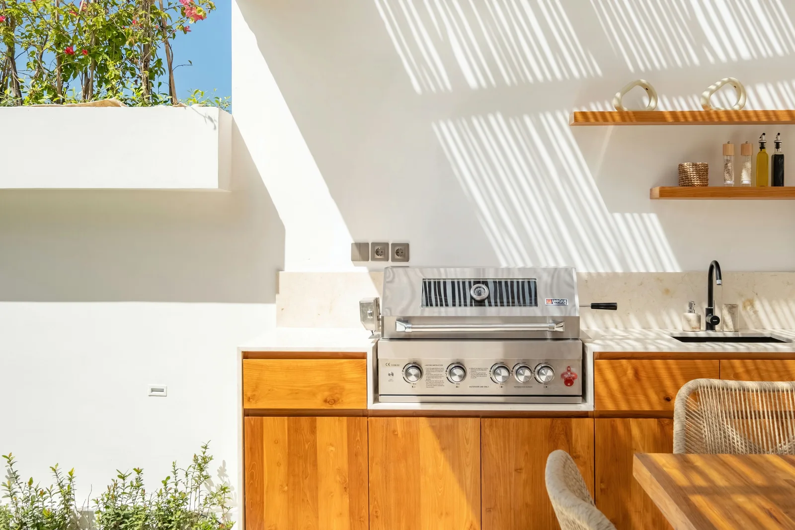 Cocina exterior luminosa con parrilla de acero inoxidable, armarios de madera y un fregadero bajo sombras rayadas.