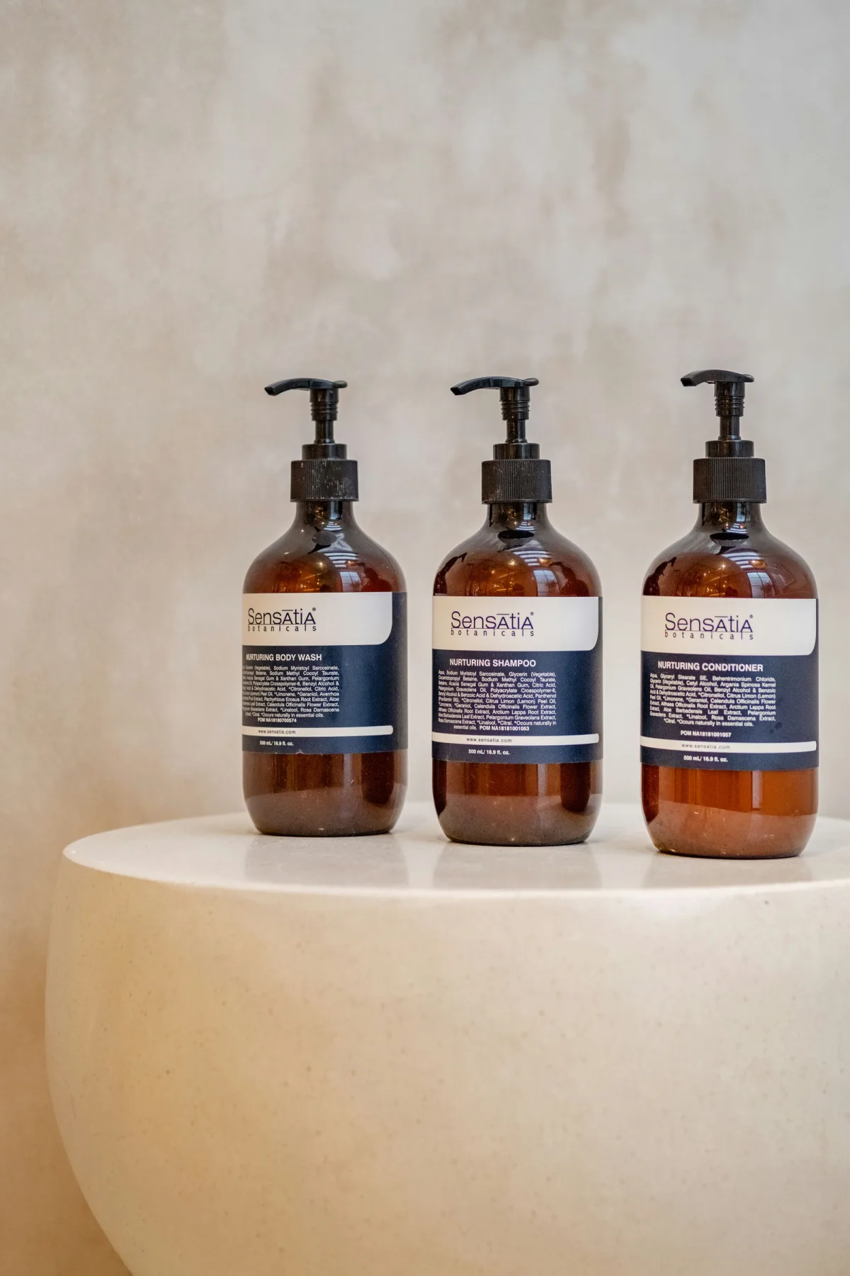 Tres botellas ámbar con dispensador están sobre un lavabo redondeado color crema, frente a una pared clara.