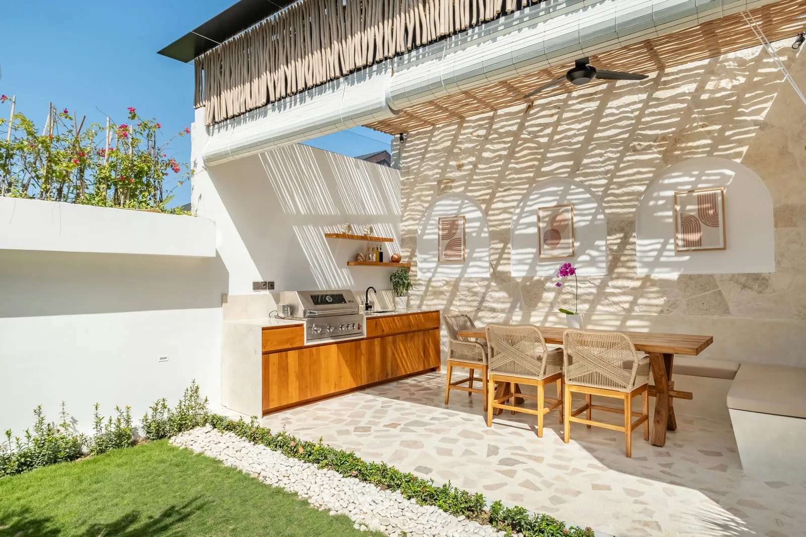 Un comedor exterior luminoso cuenta con una mesa de madera, sillas tejidas y una cocina empotrada bajo un techo de listones.
