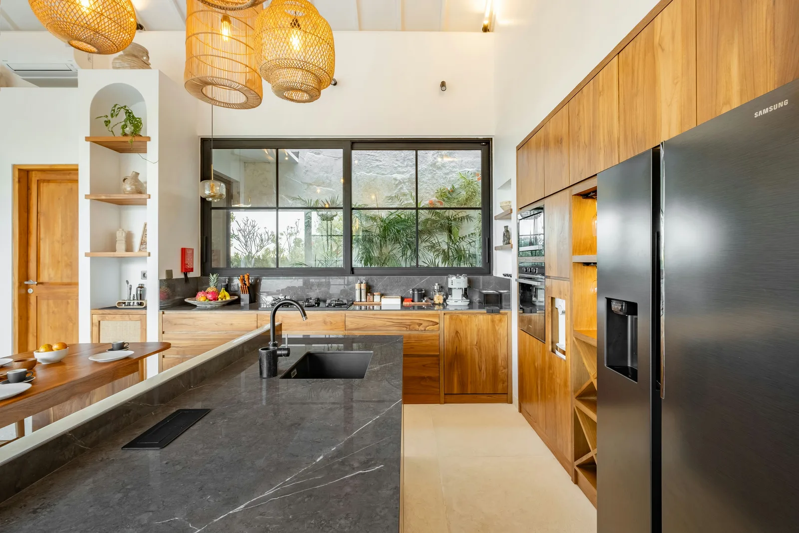 Una cocina moderna con isla de piedra oscura, gabinetes de madera, lámparas colgantes y una ventana grande.