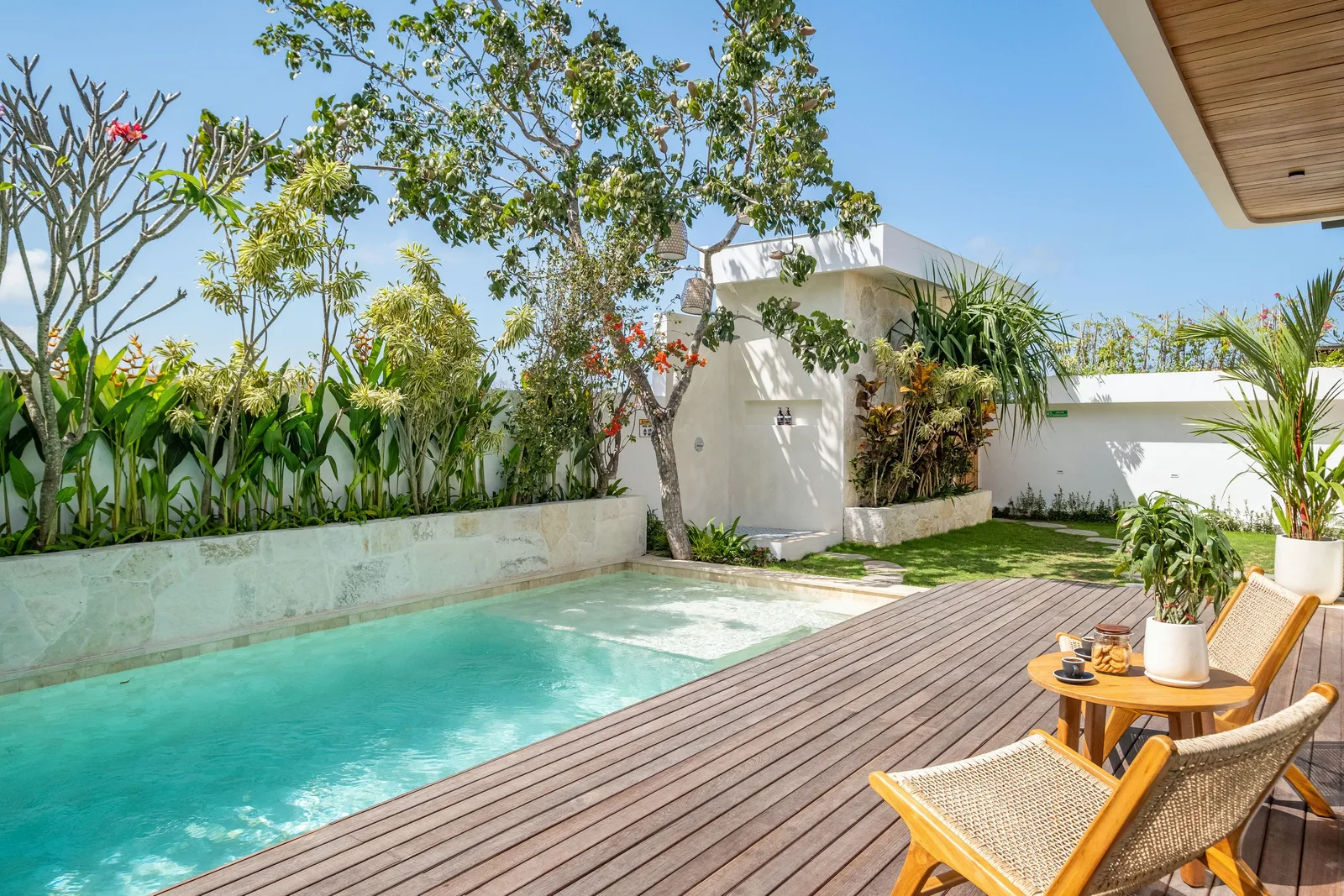 Una piscina turquesa bordea una terraza de madera con dos sillas, plantas en macetas y vegetación frondosa.