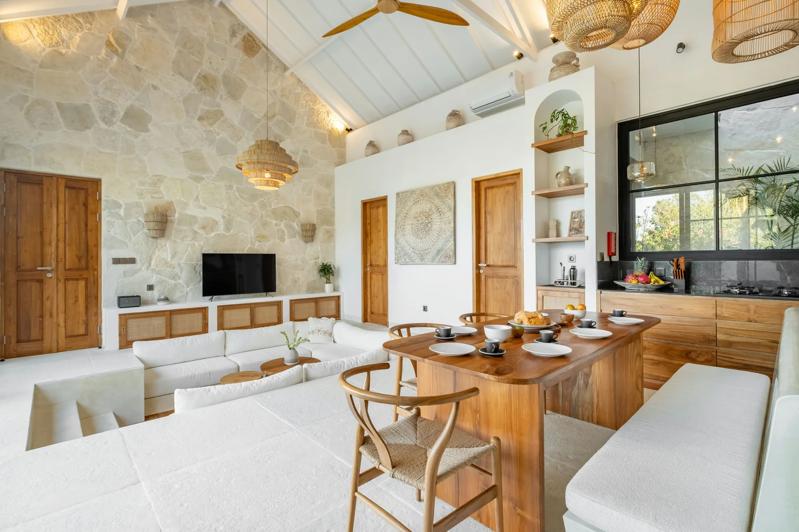 Un luminoso salón-comedor diáfano cuenta con una larga mesa de madera, sofás blancos y una pared de piedra.