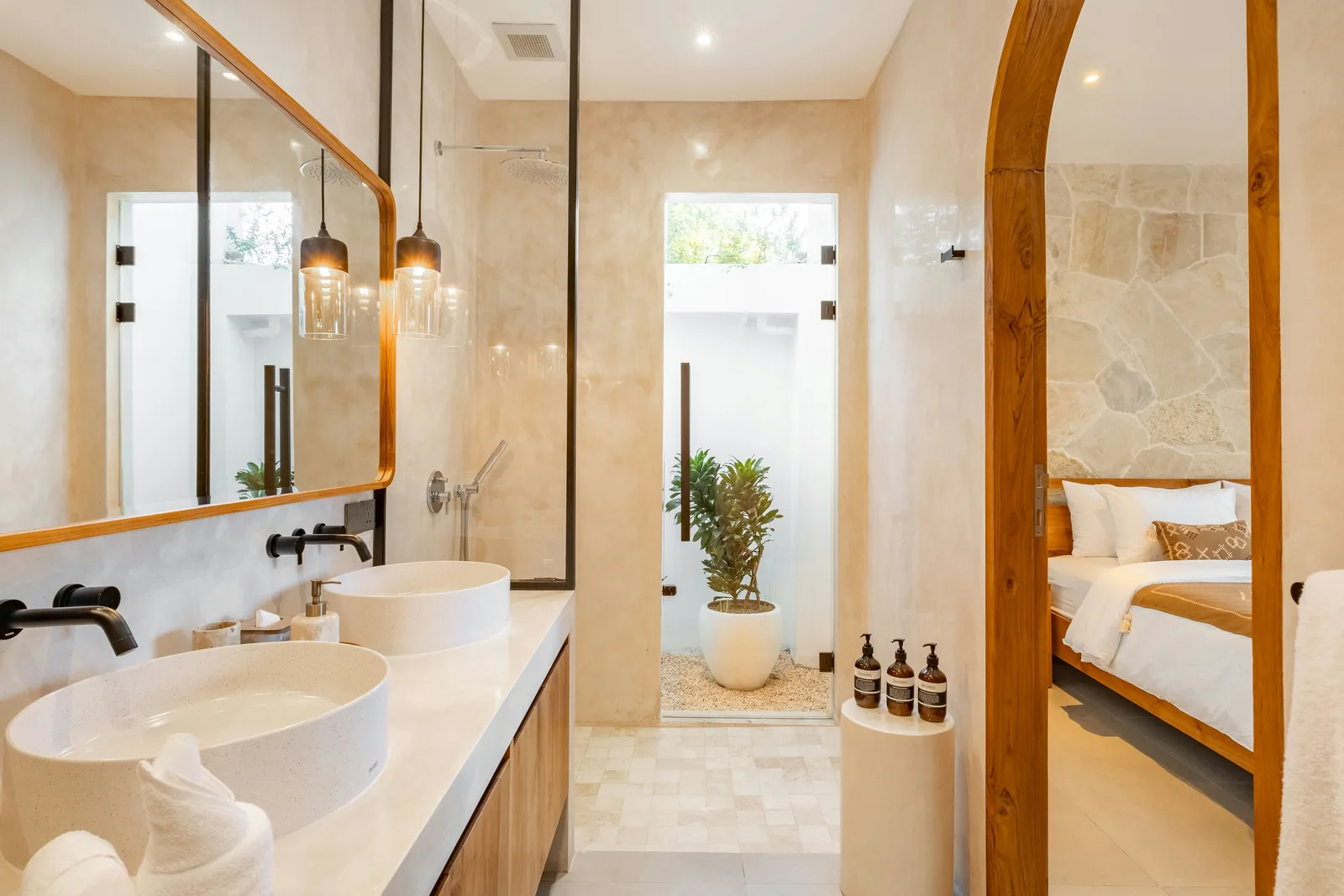 Un baño luminoso tiene dos lavabos sobre encimera, un espejo grande y una puerta hacia un dormitorio.