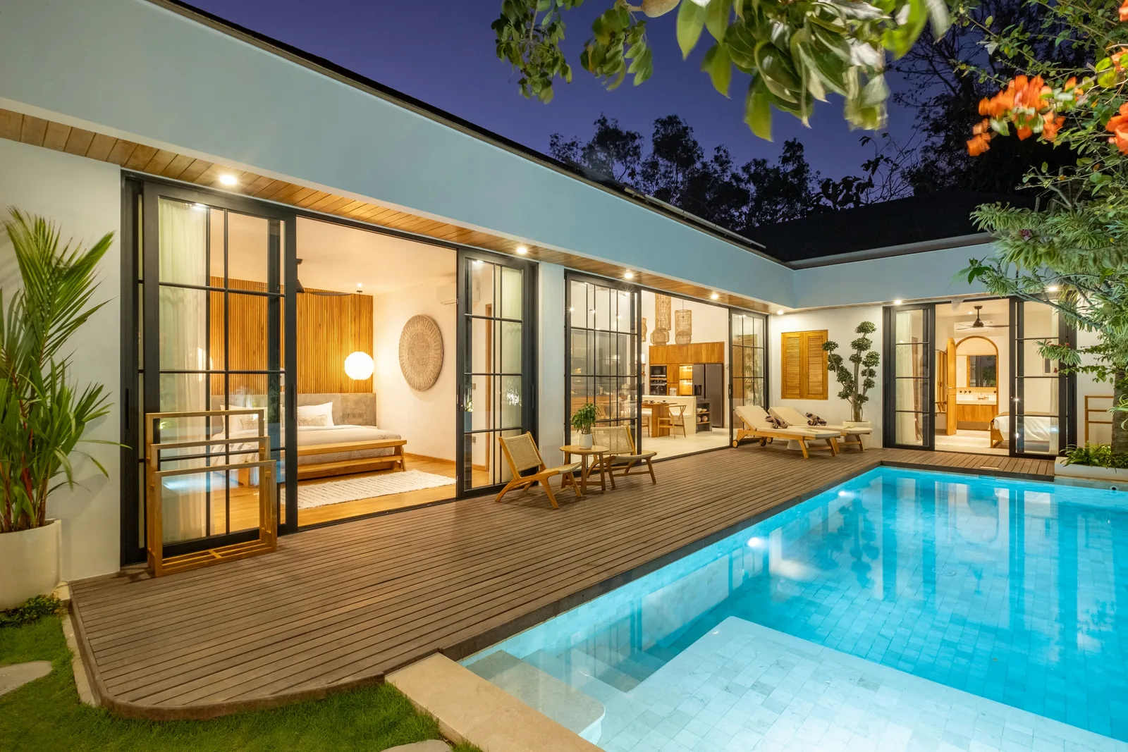Una casa moderna da a una terraza de madera con puertas de vidrio, sillas y una piscina iluminada.