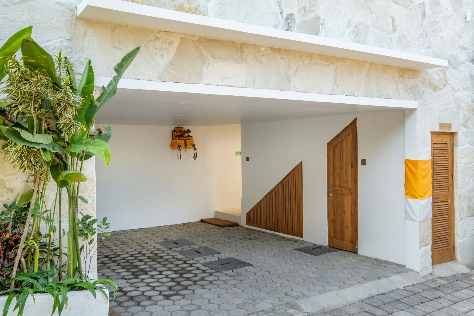 Una entrada cubierta con suelo de piedra tiene puertas de madera, un panel triangular de madera y plantas frondosas.
