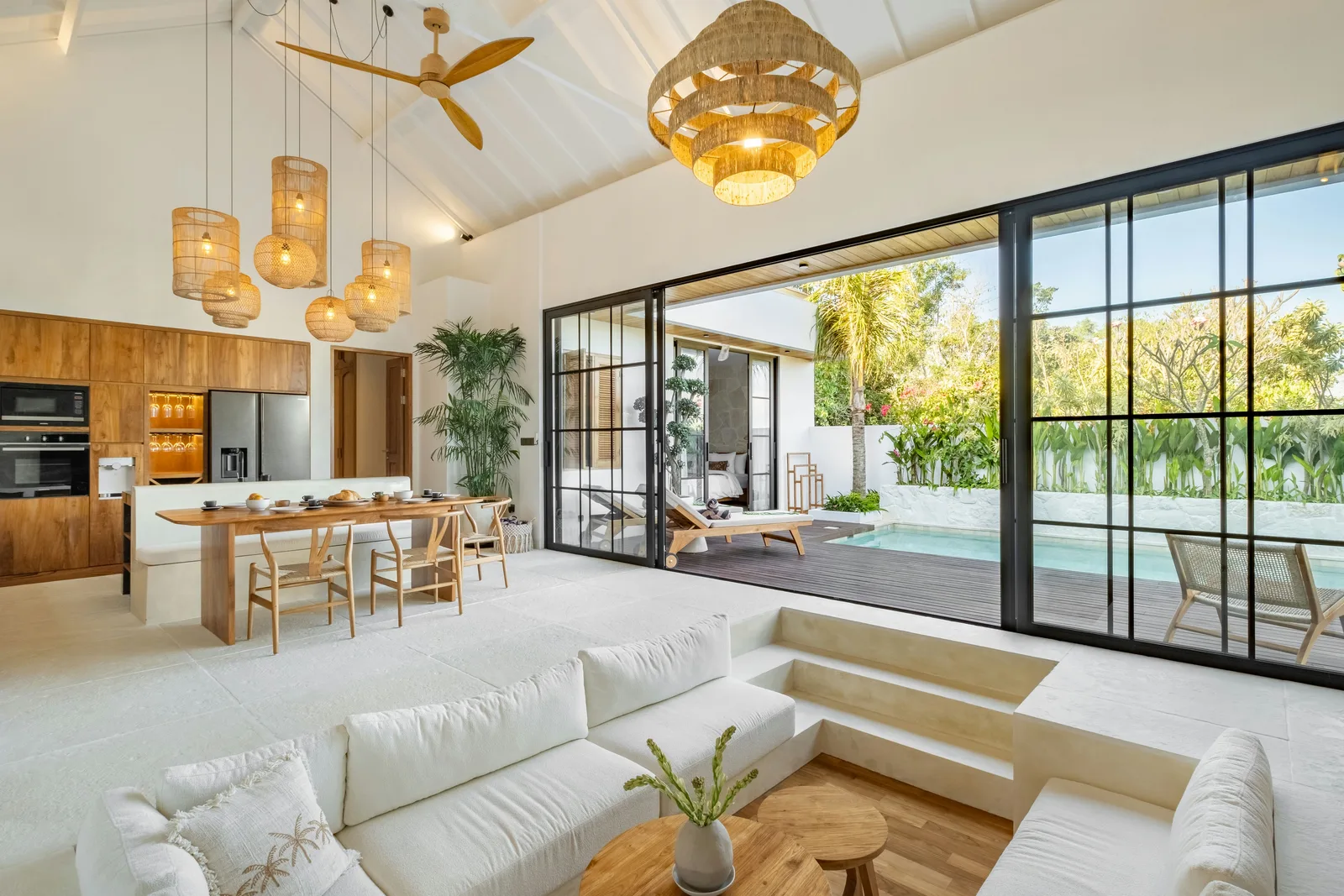 Una sala de estar y comedor de planta abierta tiene sofás blancos, una mesa de madera y puertas de vidrio que dan a un patio.