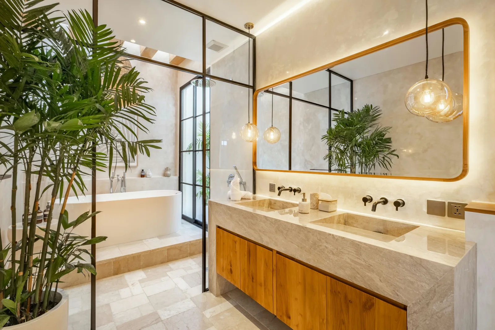 Un baño luminoso cuenta con un largo tocador de mármol, gabinetes de madera, mamparas de vidrio y una bañera exenta.