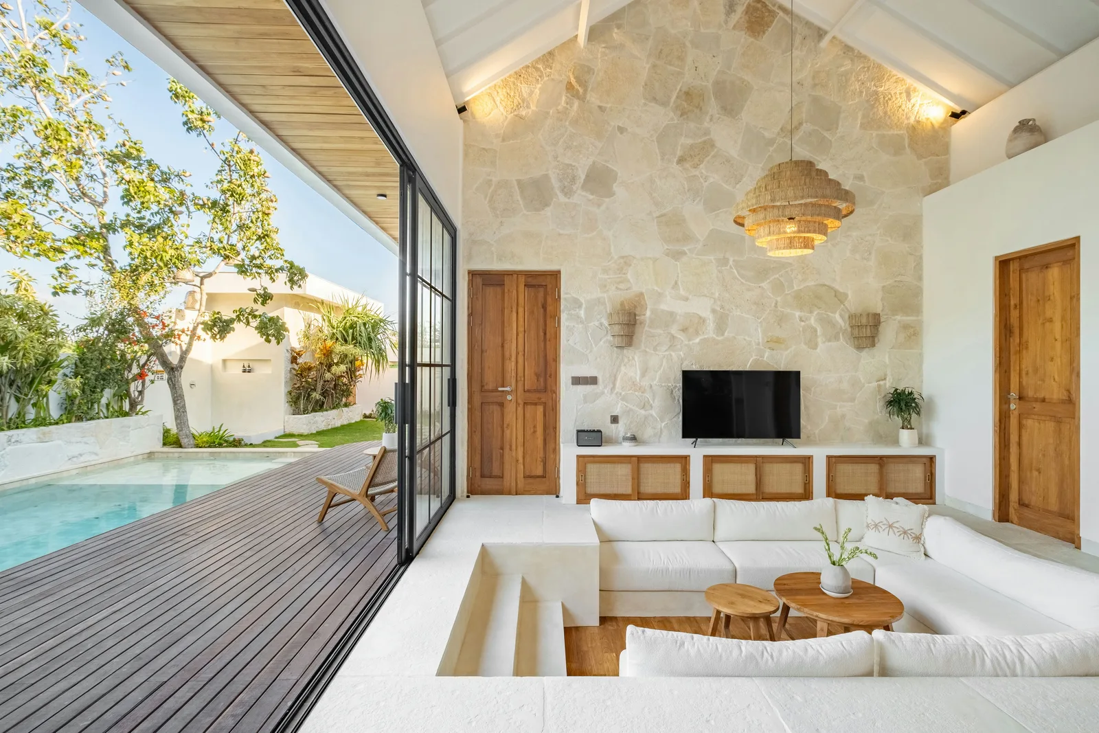 Una luminosa sala de estar se abre a una terraza de madera y una piscina, con paredes de piedra, sofá blanco y televisor.