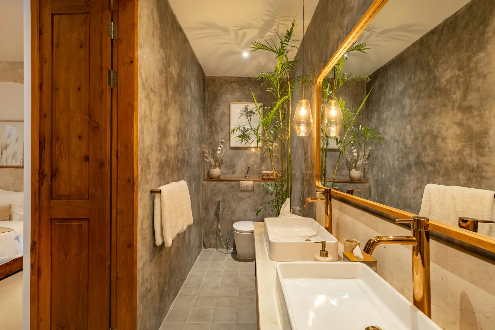 Un baño estrecho tiene dos lavabos blancos sobre encimera, un espejo con marco de madera, paredes de piedra gris y una puerta de madera.