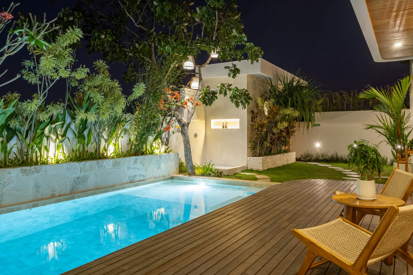 Una piscina exterior iluminada está junto a una terraza de madera, sillas de descanso y plantas tropicales por la noche.