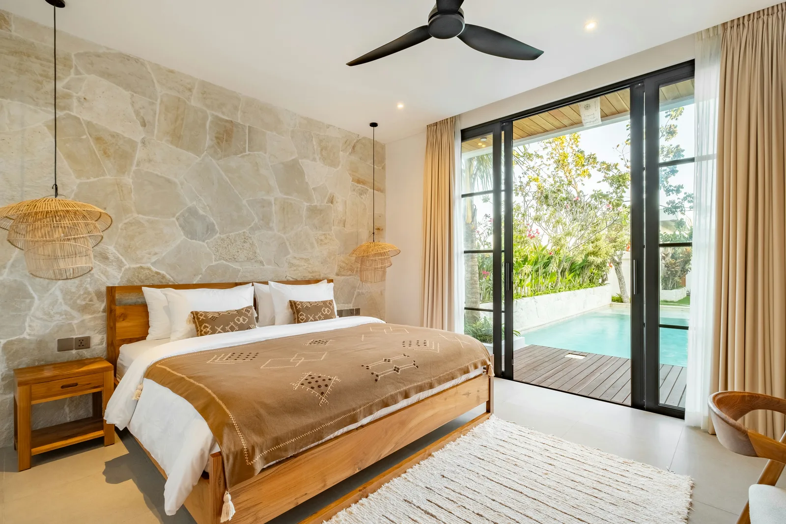Dormitorio con cama de madera, pared de piedra, ventilador de techo y puertas de vidrio con vista a la piscina.