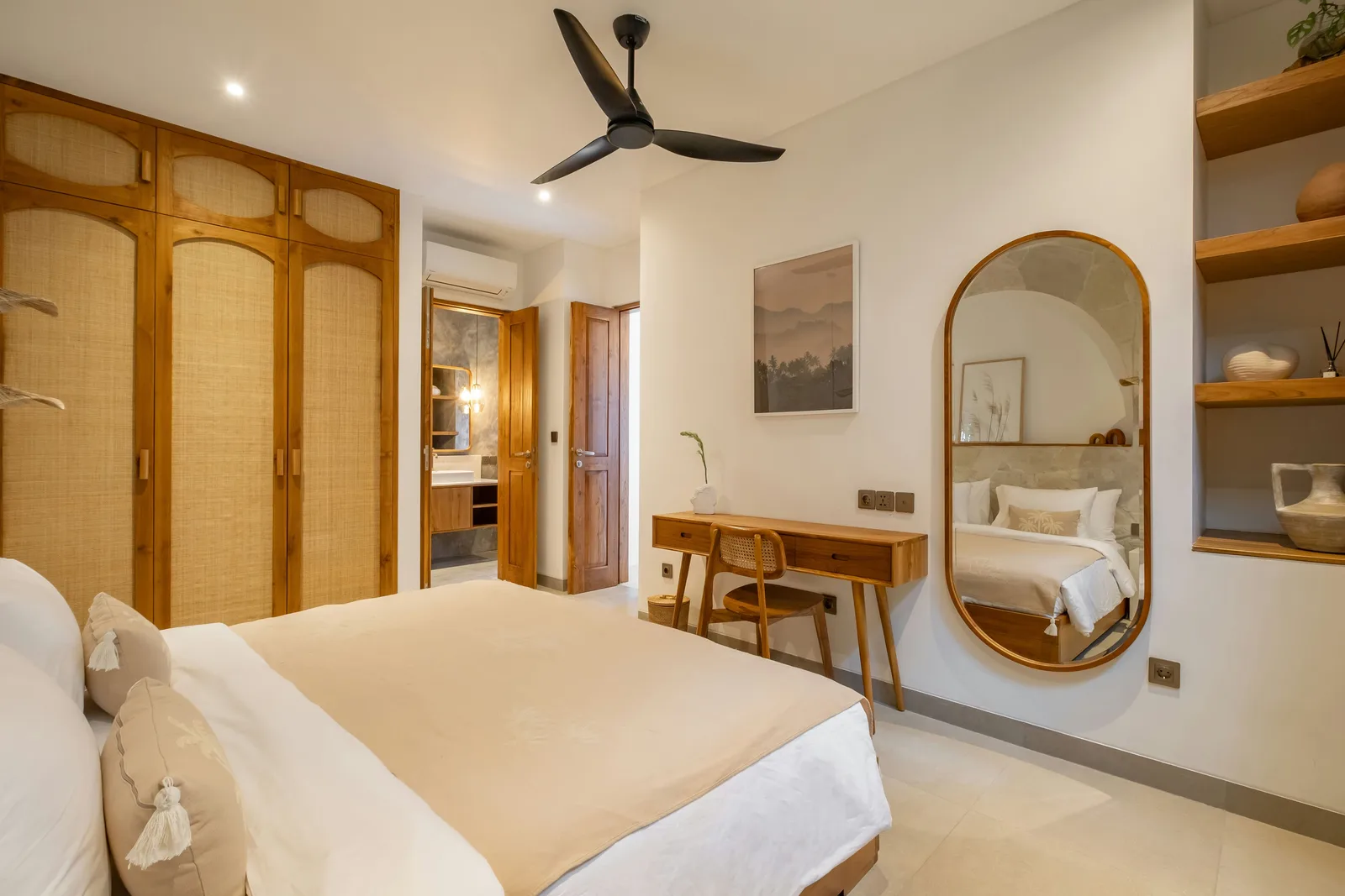 Un dormitorio luminoso tiene una cama grande, armarios con frentes de tejido, un escritorio de madera y un espejo ovalado.
