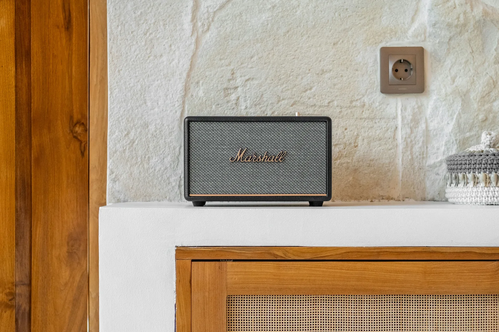 Un mueble de madera está bajo una pared de piedra blanca, junto a un pequeño altavoz y un enchufe.