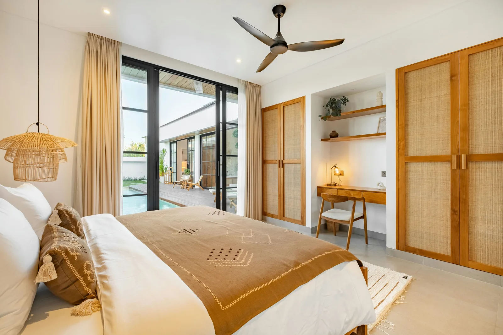 Un dormitorio luminoso tiene una cama bien hecha, una lámpara colgante de fibras y puertas de vidrio que dan a un patio.