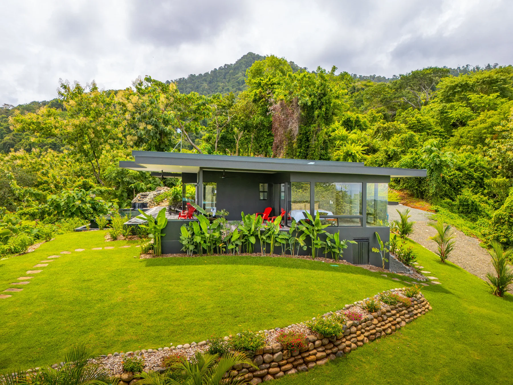 A modern pavilion sits beside a green lawn, surrounded by lush trees and hills.
