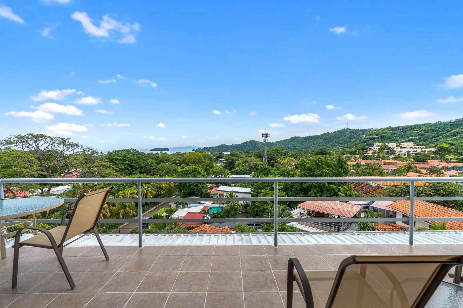 A tiled balcony with seating overlooks green hills and nearby rooftops.