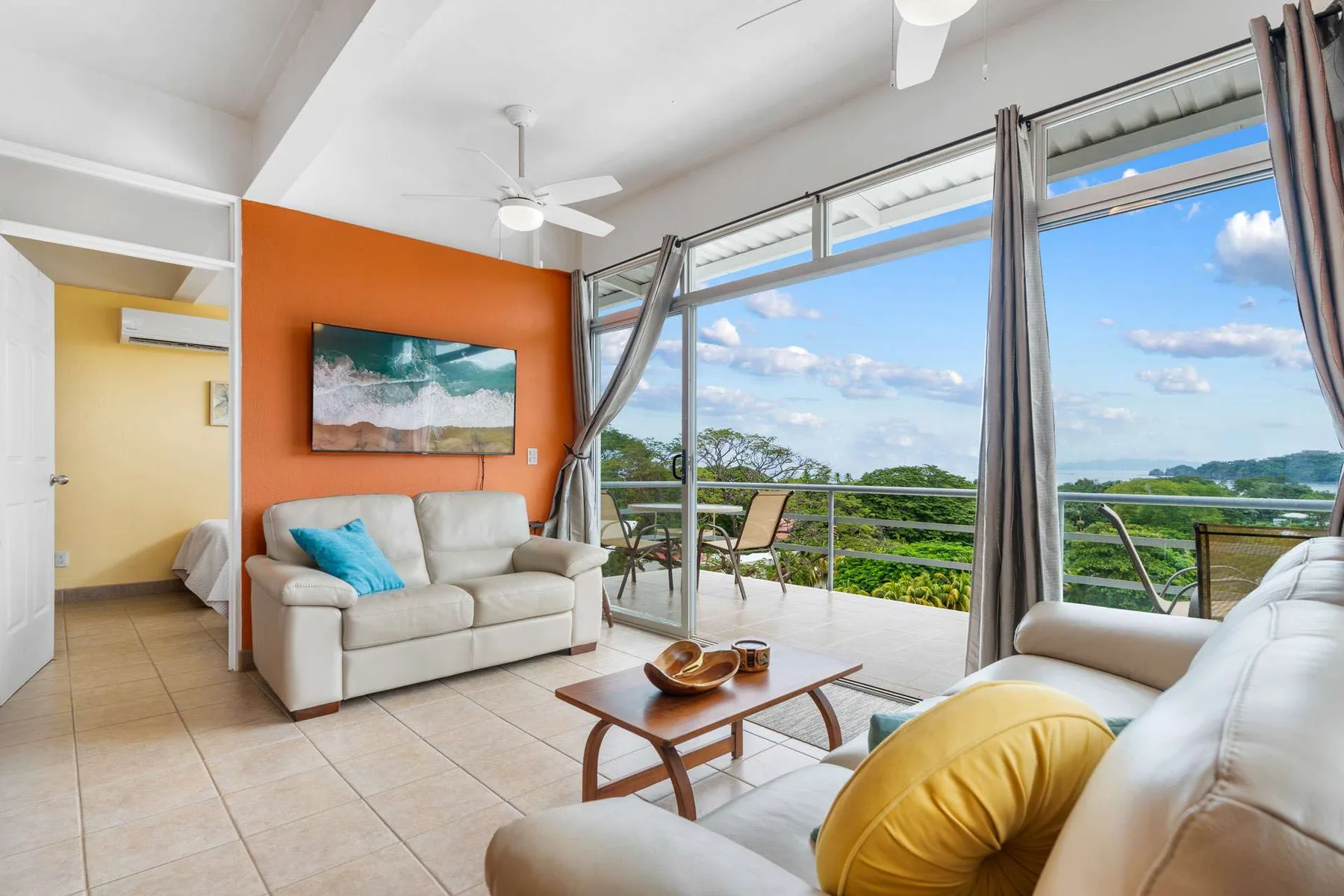 A bright living room opens onto a balcony with seating and a scenic view.