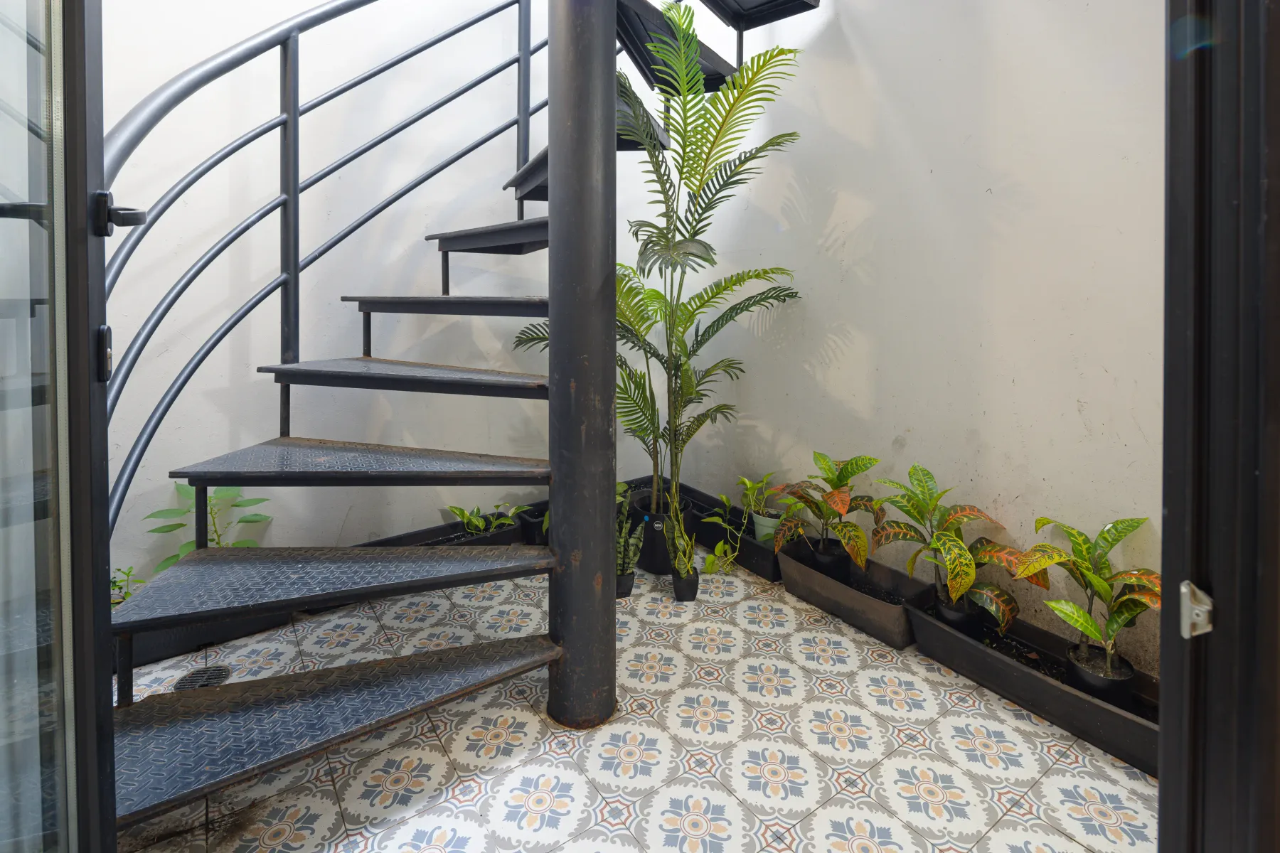 Una escalera curva de metal sube junto a plantas tropicales en macetas sobre un suelo de baldosas estampadas.
