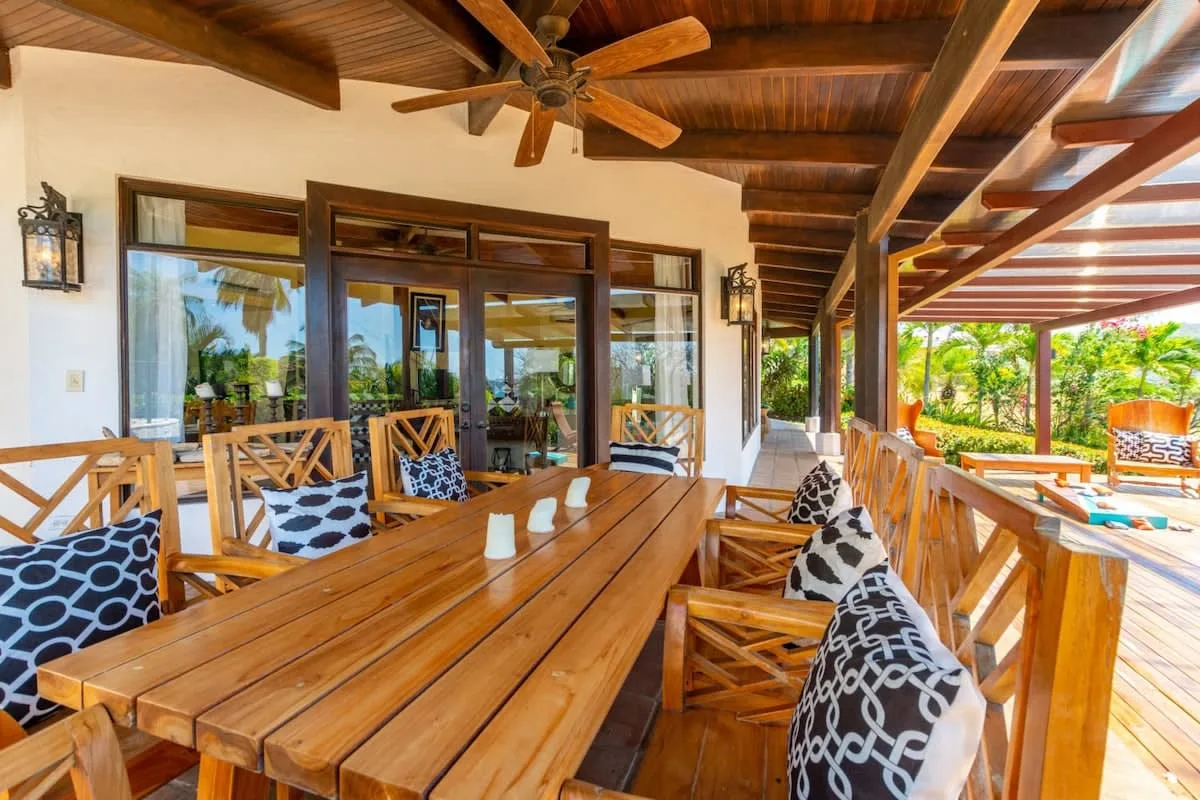 A covered wooden patio with a long dining table and seating overlooks a garden.