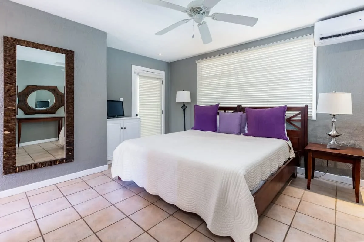 A bedroom with a bed dressed in white and purple pillows beneath a wide window.