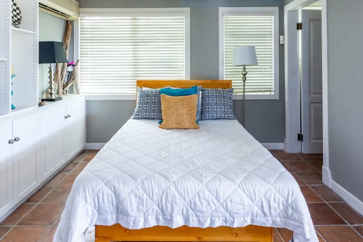 A bedroom with a bed dressed in white bedding and windows with blinds.