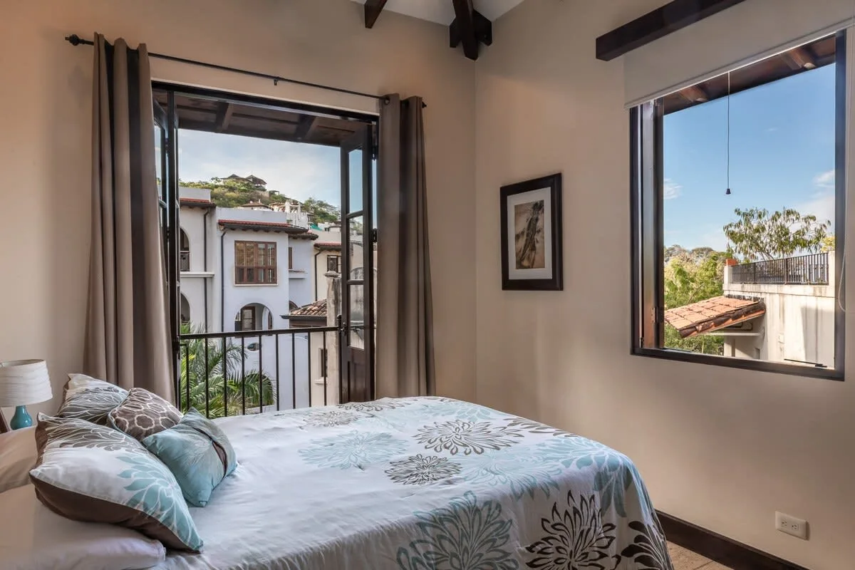 Un dormitorio con una cama junto a dos grandes ventanas con vistas a edificios y vegetación cercanos.
