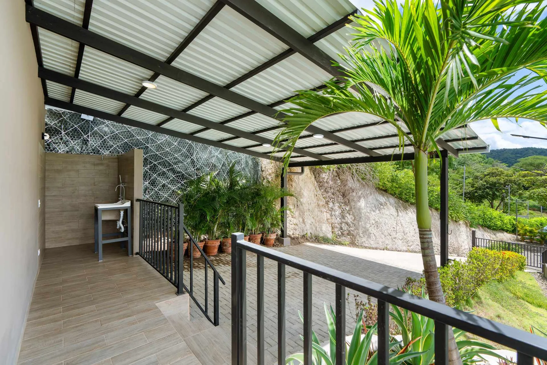 A covered outdoor terrace with a sink, railing, and palm tree.