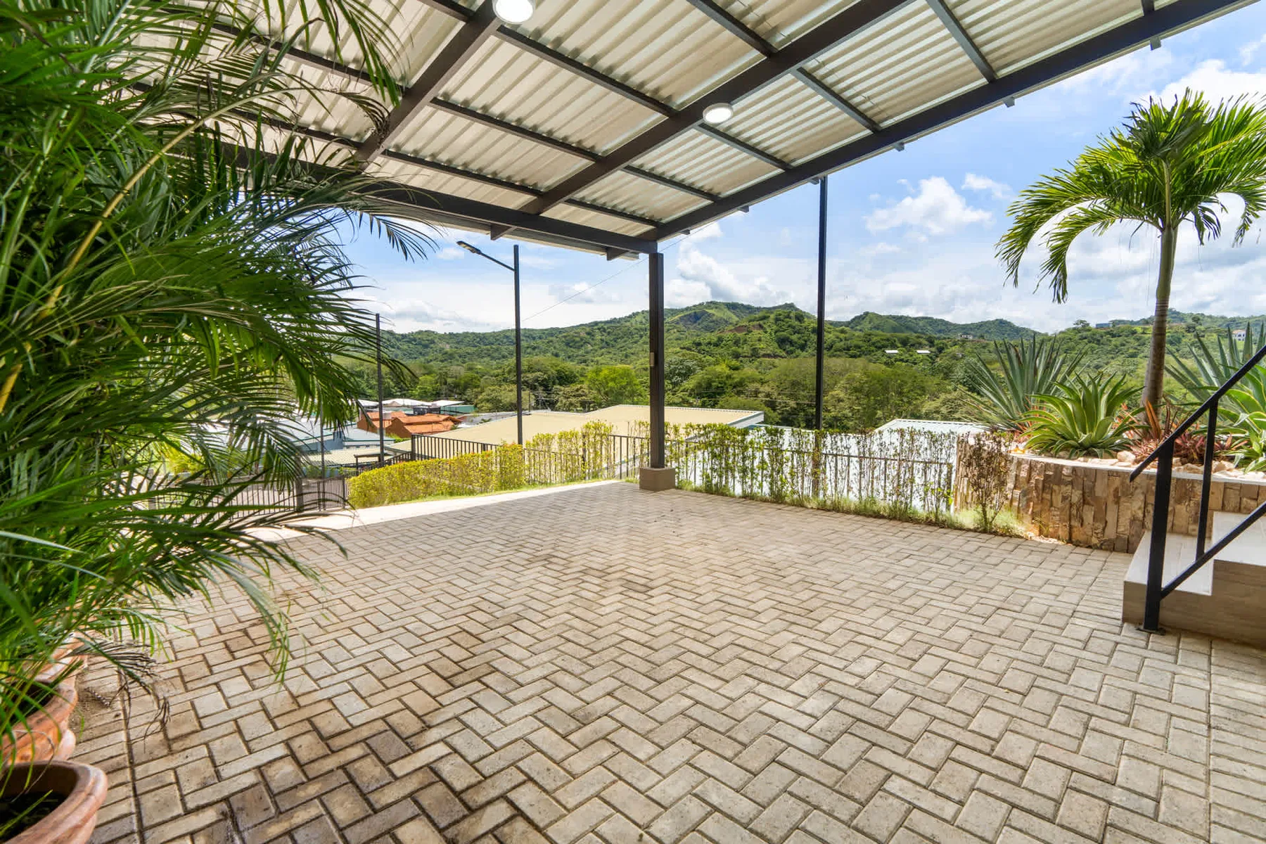 A covered patio with paved flooring overlooks green hills and surrounding vegetation.