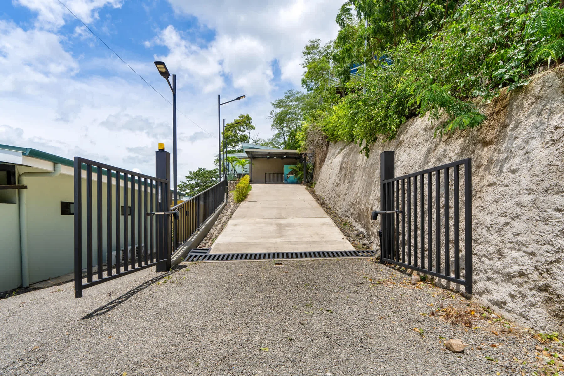 A paved driveway passes through an open metal gate beside a rocky, tree-lined slope.