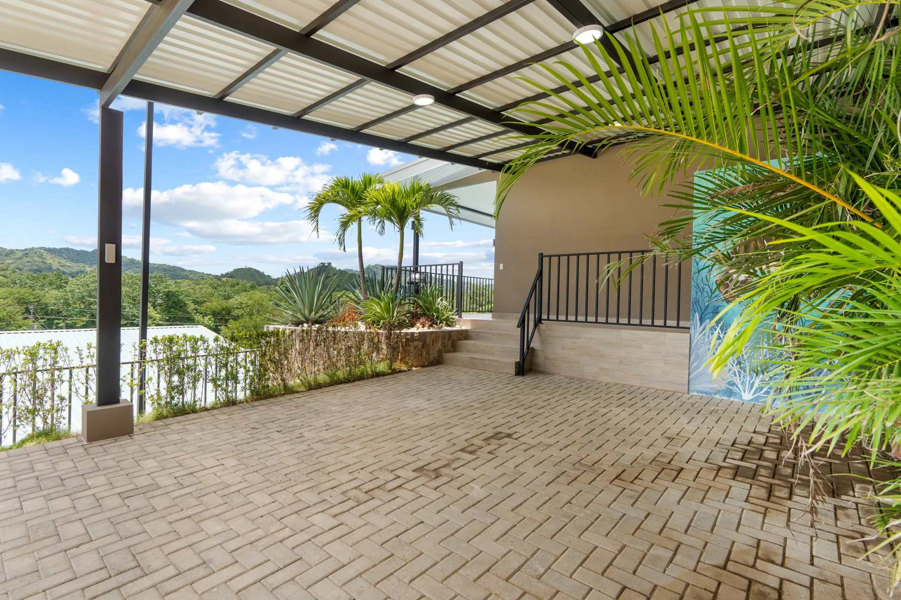 A covered outdoor terrace with paved flooring, railings, and surrounding greenery.