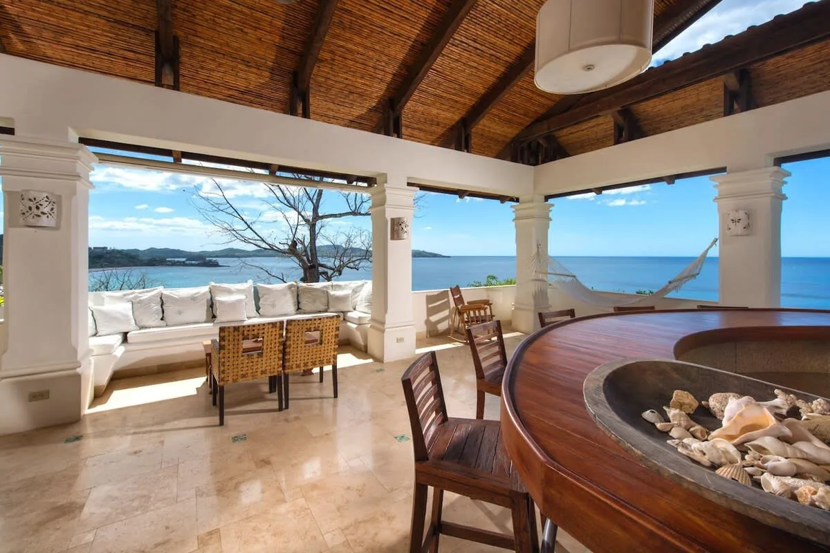 An open-air dining area overlooks the ocean, with a large round wooden table.