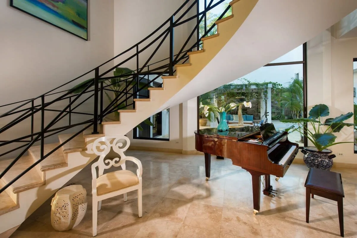 A bright interior with a staircase and a grand piano beside large windows.