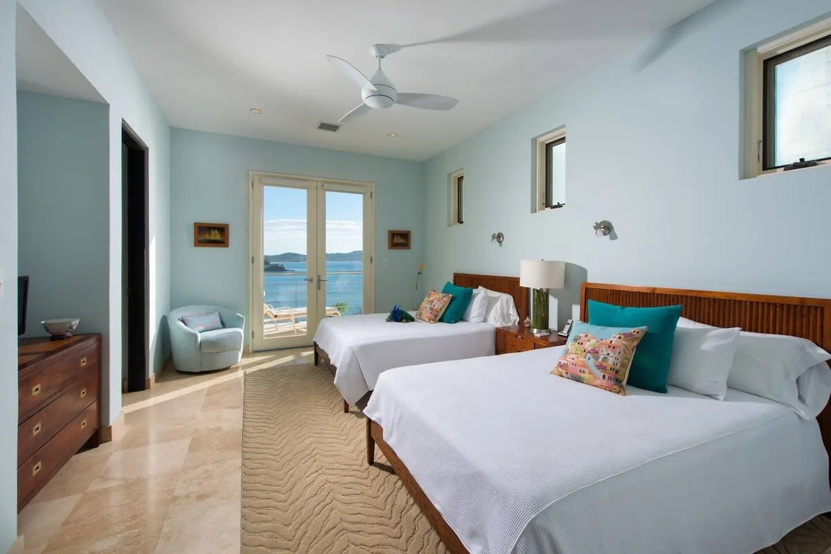 A bright bedroom with two beds and glass doors overlooking the water.