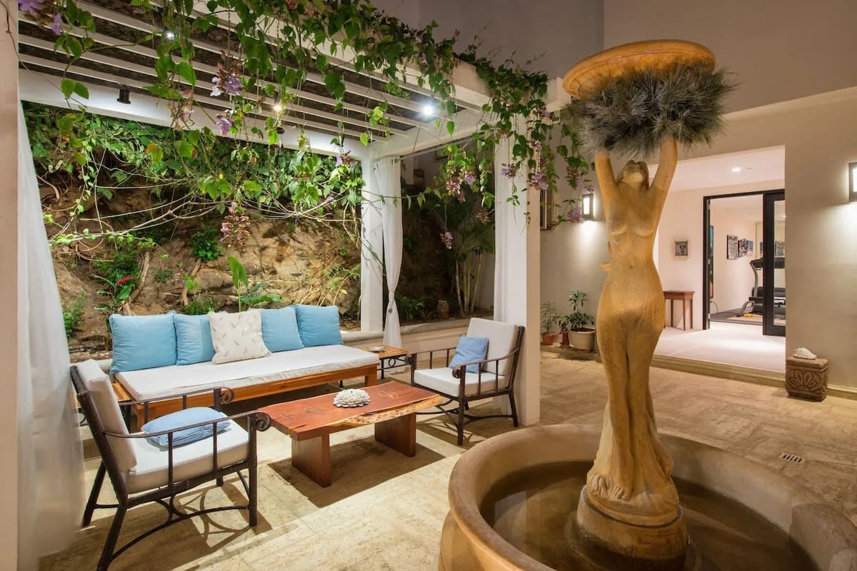 A patio seating area beneath a vine-covered pergola faces a sculptural fountain.