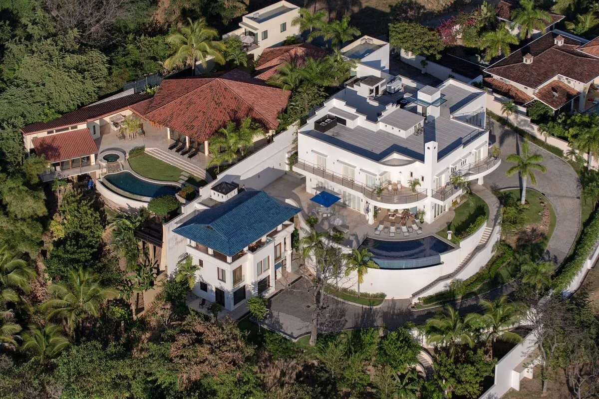 An aerial view shows a modern white building and neighboring homes among dense greenery.