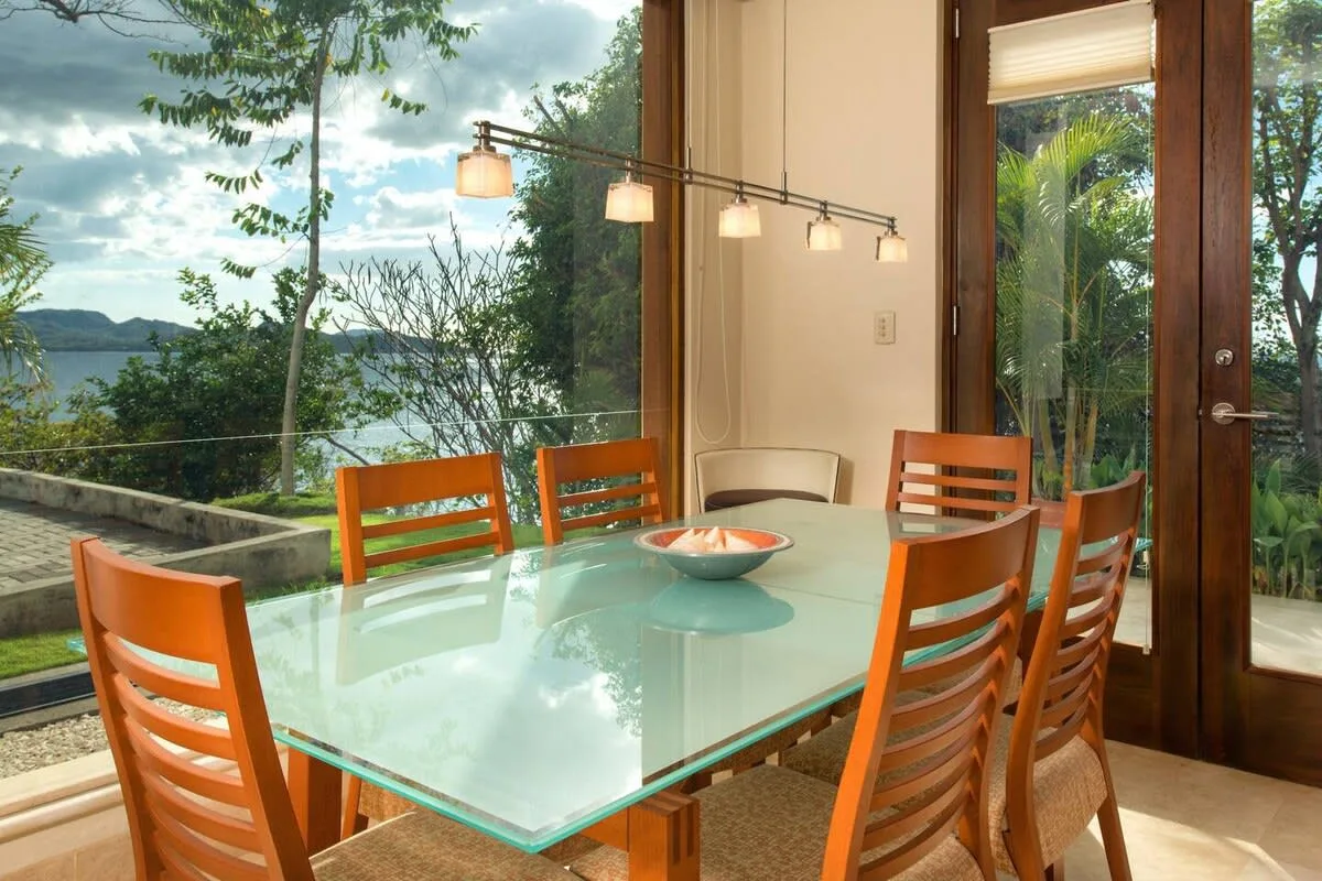 A dining area with a glass table and wooden chairs beside large windows.