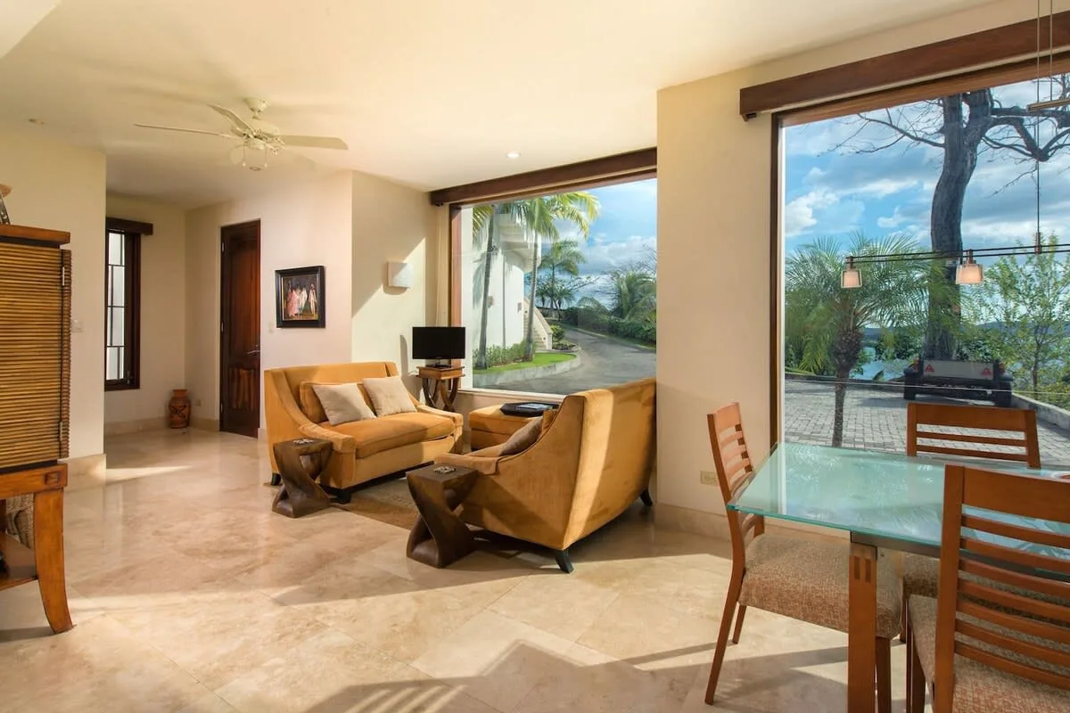 A living room with seating beside a glass dining table and large windows.