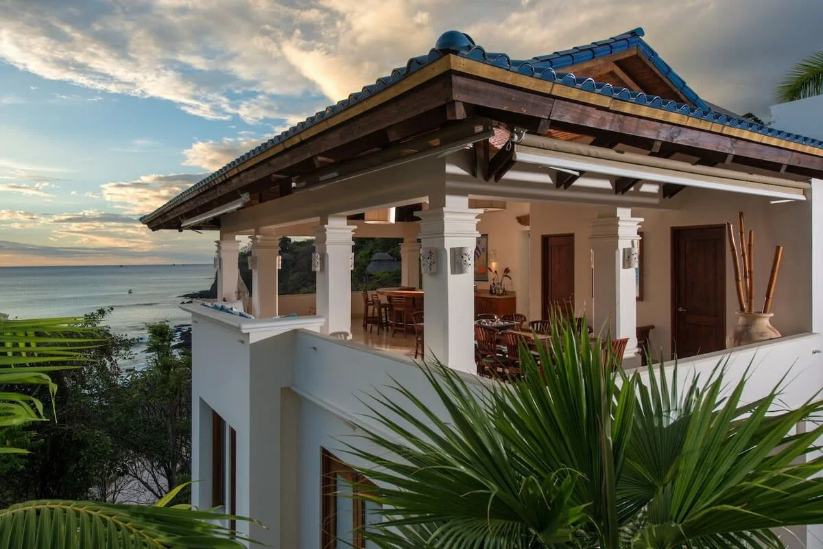 A seaside house has a covered terrace with white columns and outdoor seating.